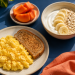Ovos mexidos com pão, iogurte com aveia e banana e mamão sobre uma mesa azul.
